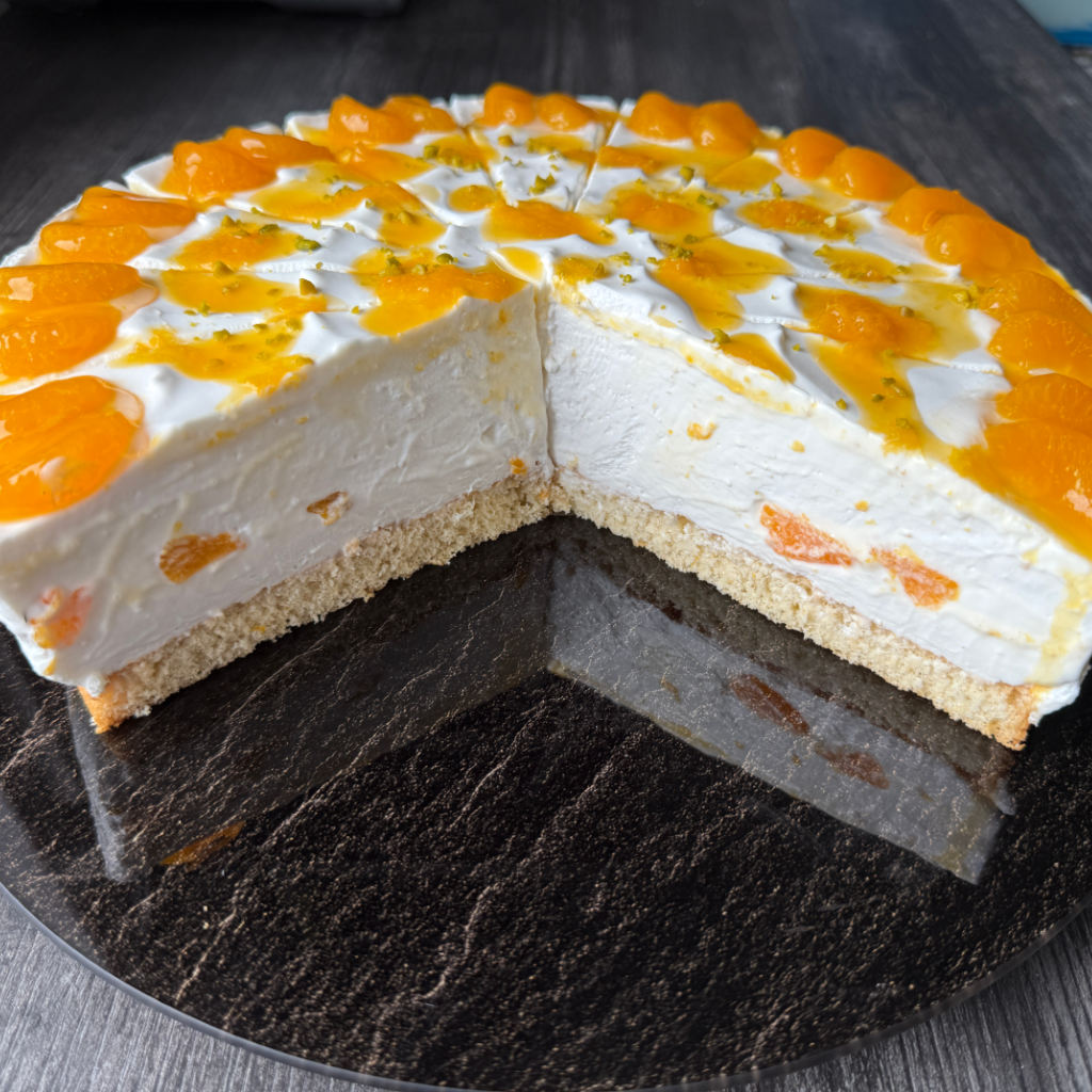 Eine angeschnittene Mandarinen-Mascarpone-Torte mit hellen Cremeschichten, Mandarinen-Deko und gehackten Pistazien.