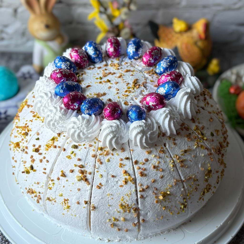 Festlich dekorierte Osterkuppeltorte mit weißer Sahnehaube, Pistazien, Krokant und bunten Schokoeiern auf einer Tortenplatte.