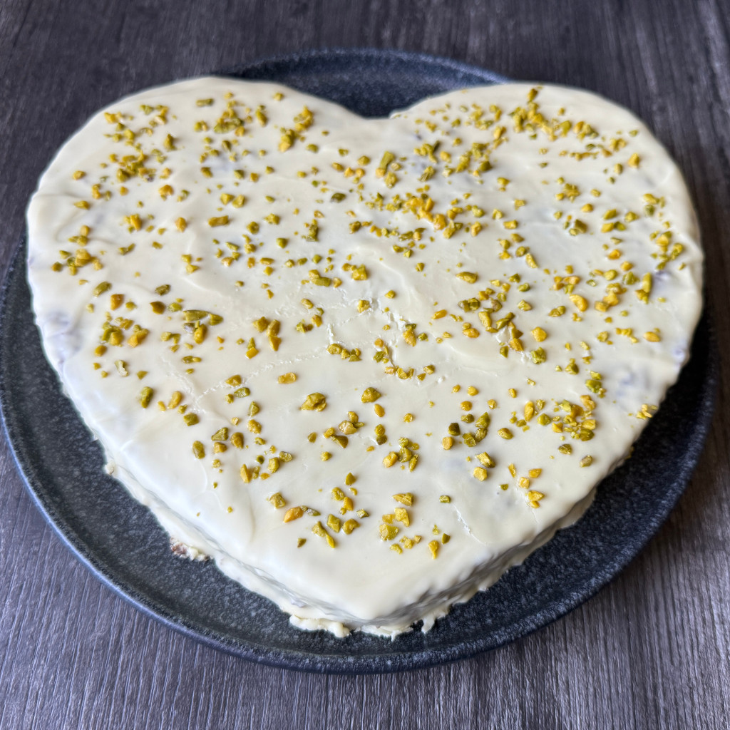 Herzförmiger Schokoladenkuchen mit weißem Schokoladenüberzug und grünen Pistazien auf einer Tortenplatte.
