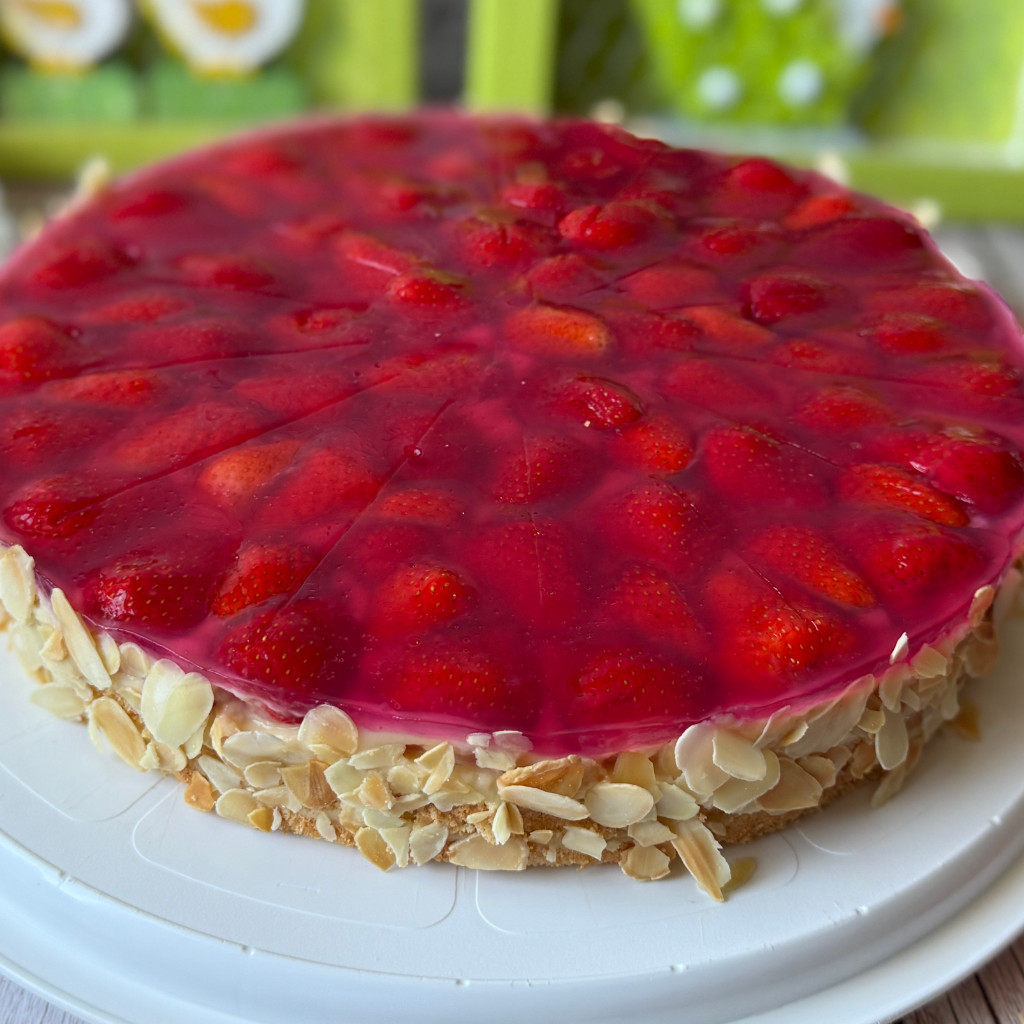 Ein fertig dekorierter Erdbeer-Pudding-Kuchen auf einer Tortenplatte. Die Oberfläche ist dicht mit glänzenden, roten Erdbeerhälften und Tortenguss belegt. Der Kuchenrand ist gleichmäßig mit goldgelb gerösteten Mandelblättchen bedeckt.